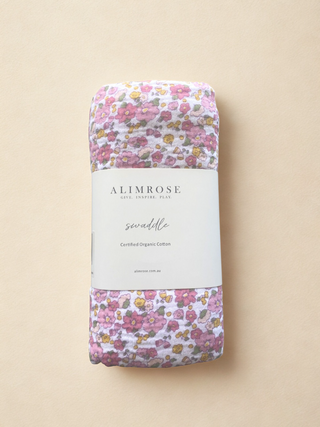 Floral swaddle blanket with 'Alimrose' card branding wrapped around it on a beige background. Fabric is white with light pink, dark pink, yellow and cream flowers and green leaves.