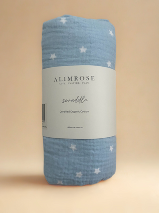Blue swaddle blanket with a white star pattern, pictured standing up (rolled) against a beige background. White Alimrose branded card wrapped around it. 