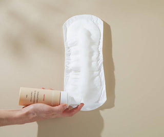 Hand holding a Bare Mum perineal foam and pressing it down across a sanitary pad. Pictured on a beige background.